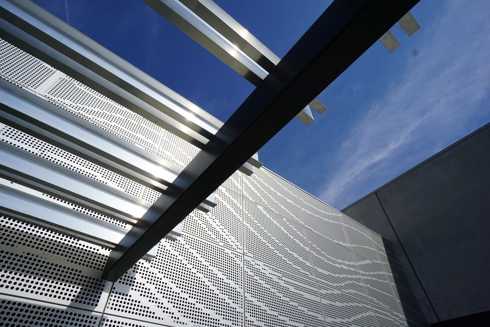 Perforated metal facade and steel pergola structure on a modern building exterior against a blue sky.
