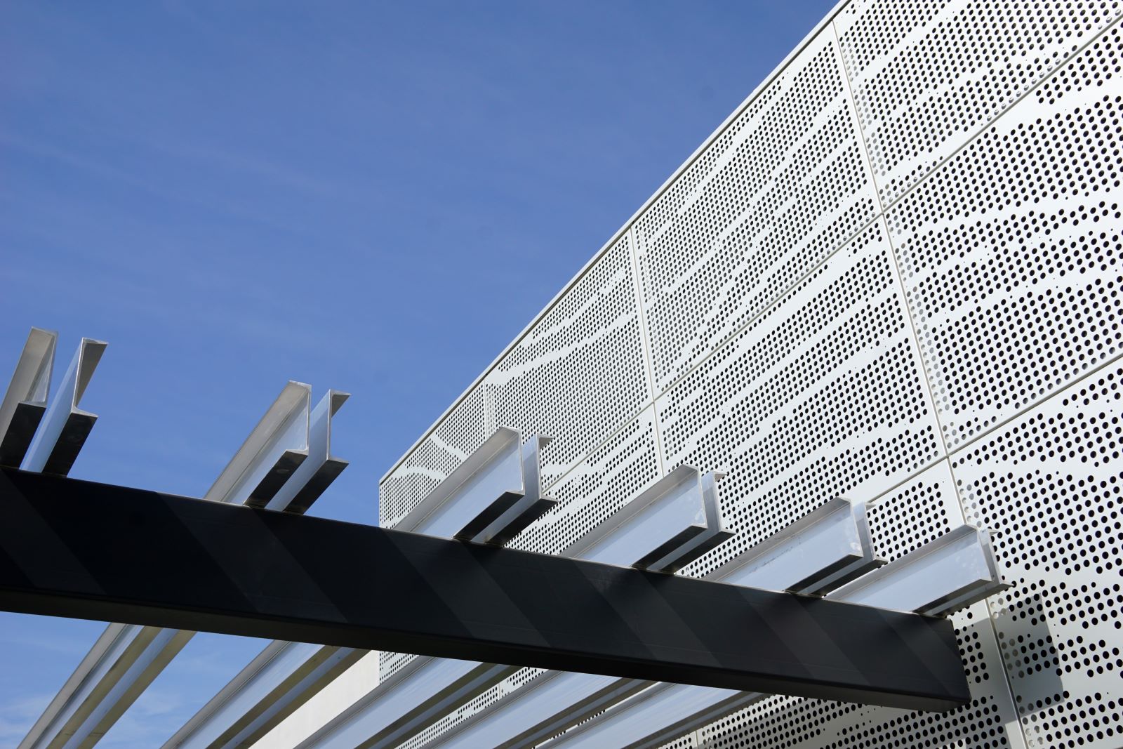 Perforated metal facade and steel beam architectural details against a clear blue sky.