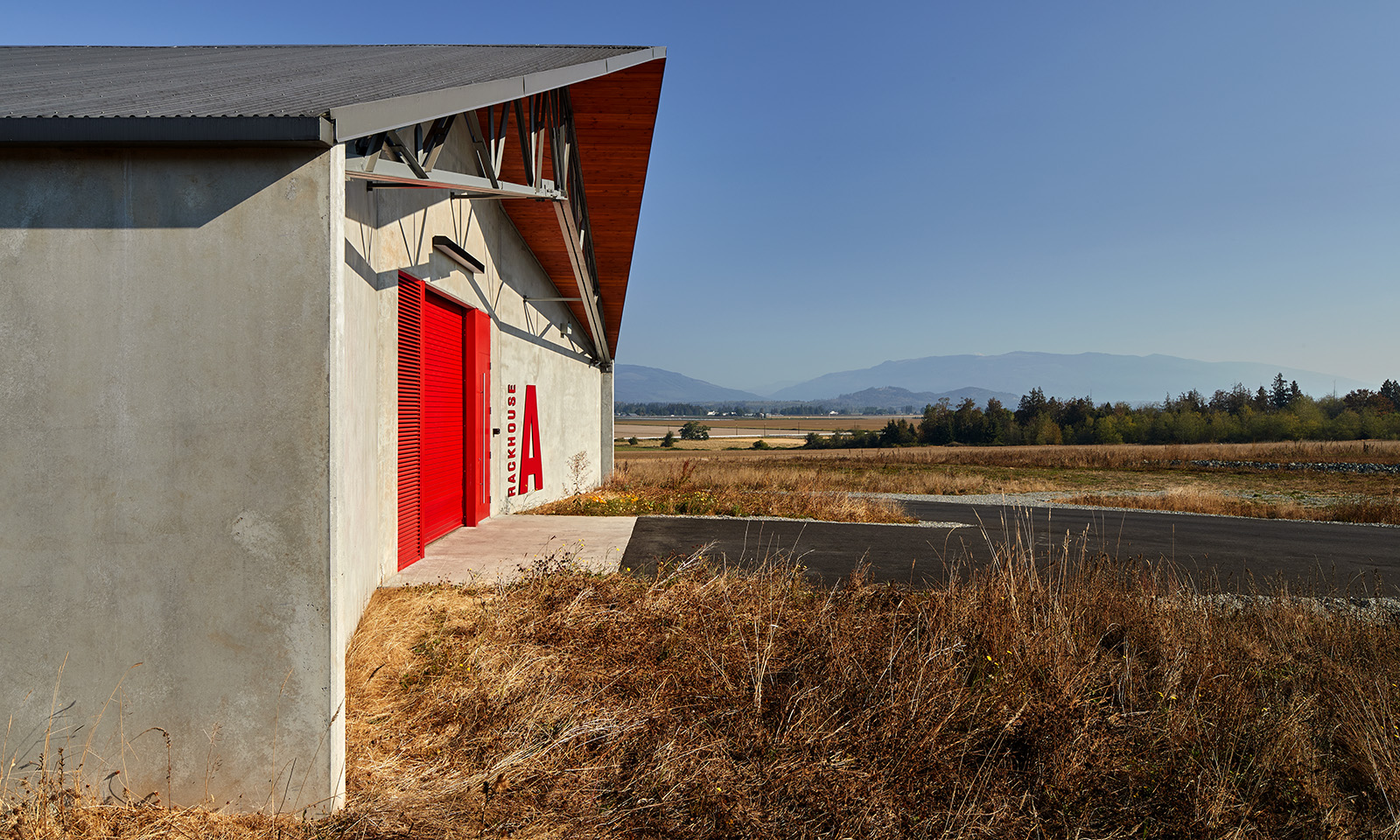 Modern concrete rackhouse with a bright red door, set in a dry grassy field with distant mountains.