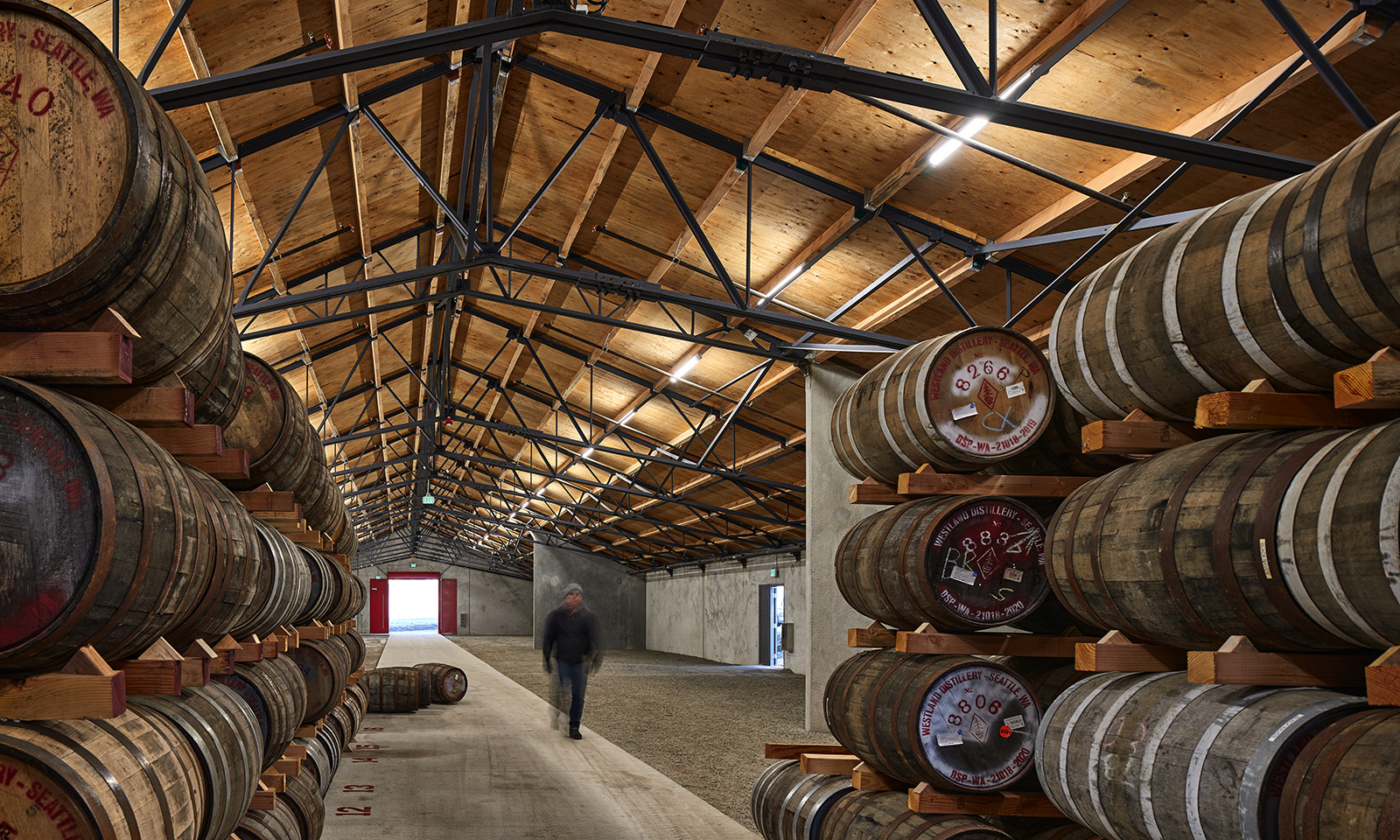 Rows of wooden barrels stacked in a spacious rackhouse with exposed timber ceilings and steel trusses.