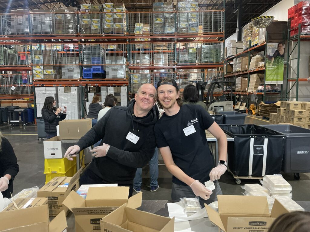 Two volunteers smiling while packing boxes in a large food bank warehouse.