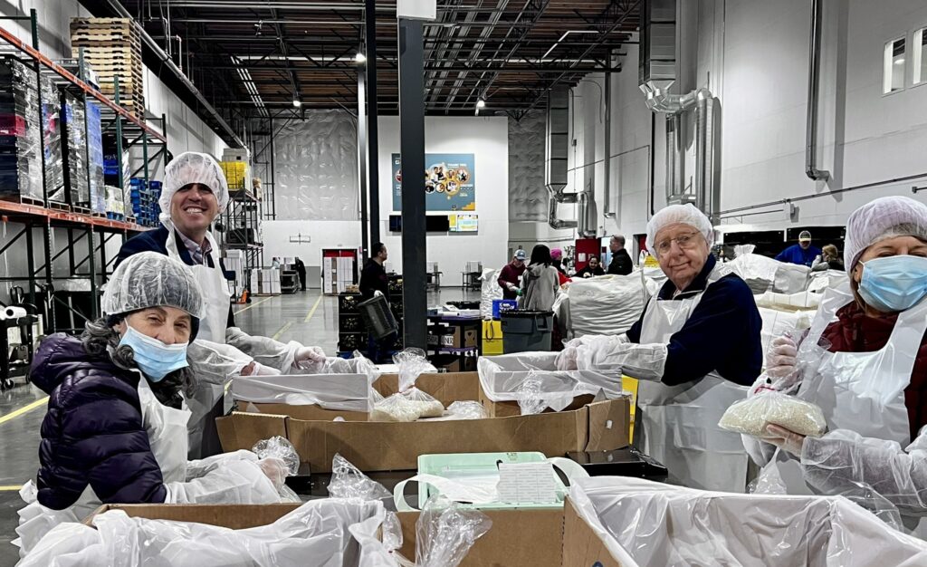 Volunteers wearing hairnets and aprons pack bags of food into boxes inside a large warehouse.