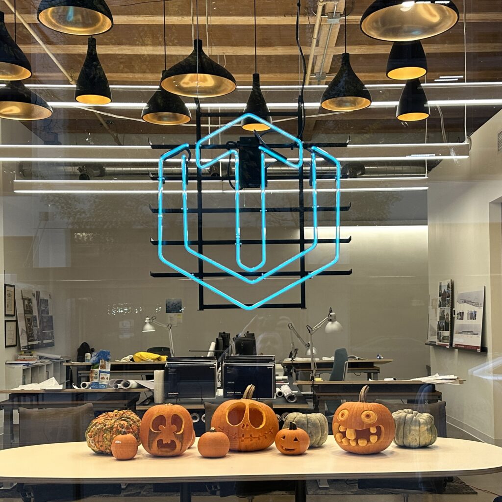 Carved jack-o'-lanterns displayed on a table inside an office window under a glowing blue neon sign.