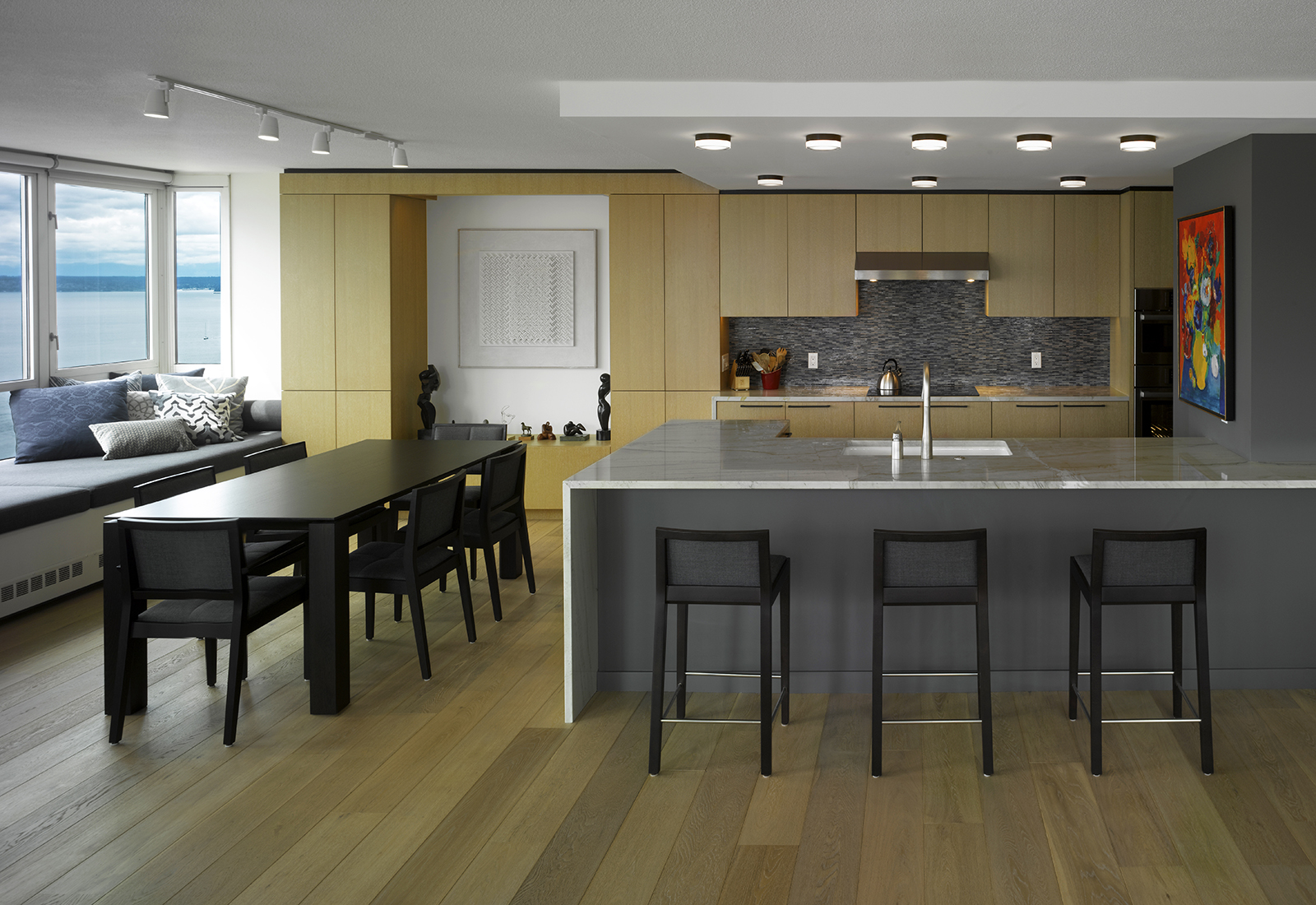 Modern kitchen and dining space with light wood cabinets, a grey island, black dining set, and scenic water views.