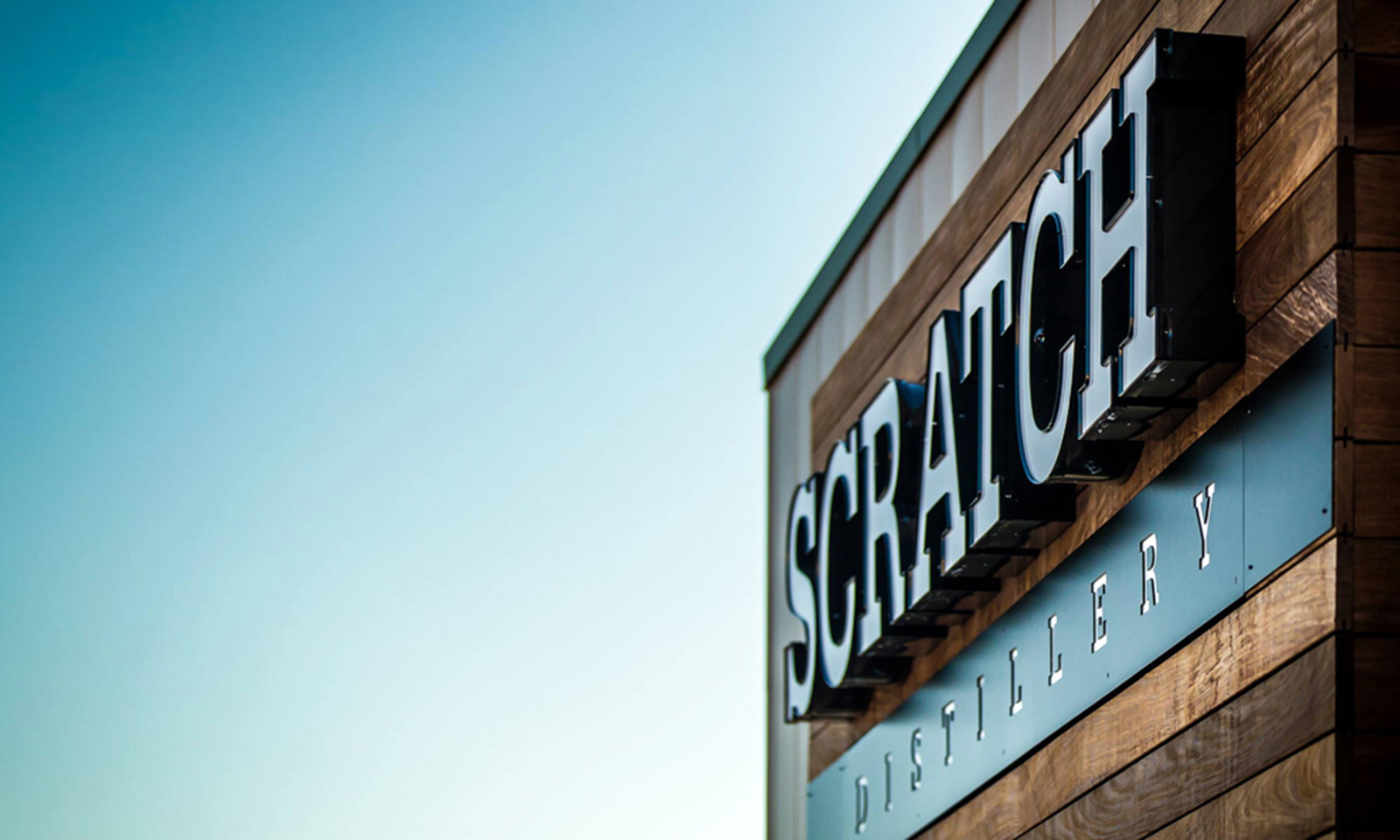 Dimensional Scratch Distillery sign mounted on horizontal wood siding against a clear blue sky.