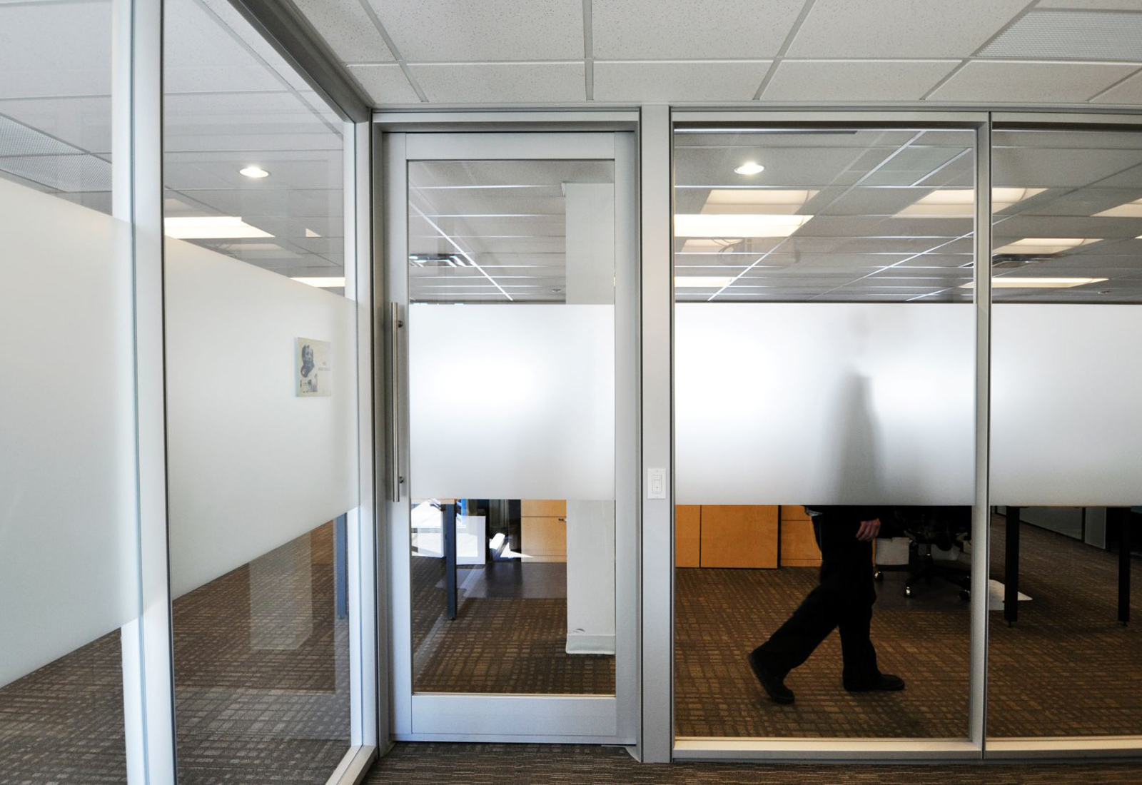 Modern office interior with aluminum-framed glass walls, frosted privacy film, and a person walking behind.