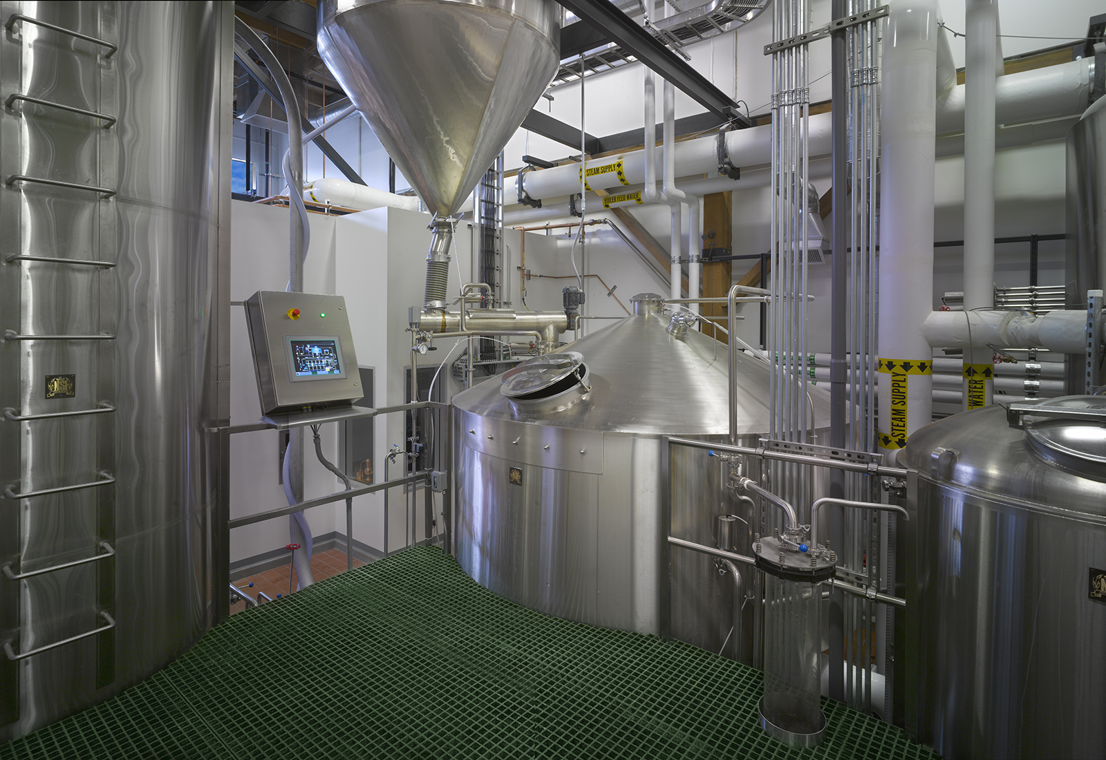 Industrial distillery interior featuring stainless steel mash tuns, overhead piping, and green grated flooring.