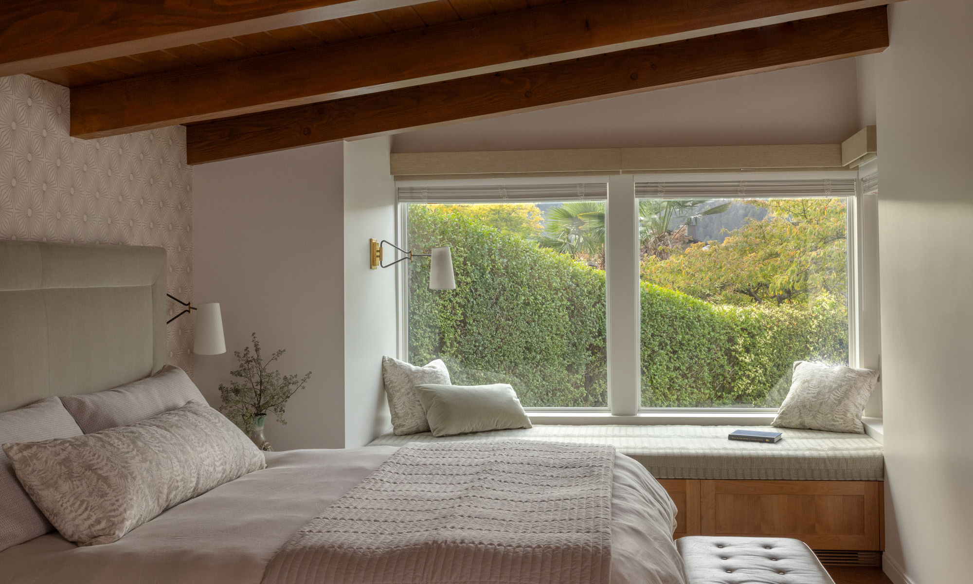 Bedroom with exposed wood beams, neutral bedding, and a cozy window seat looking out at lush green hedges.