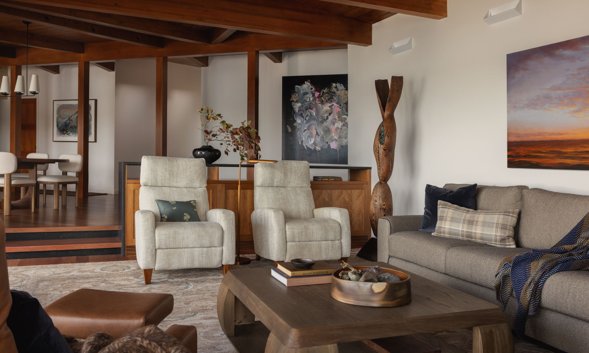 Cozy mid-century modern living room with exposed wood ceiling beams, a grey sofa, and two light armchairs.