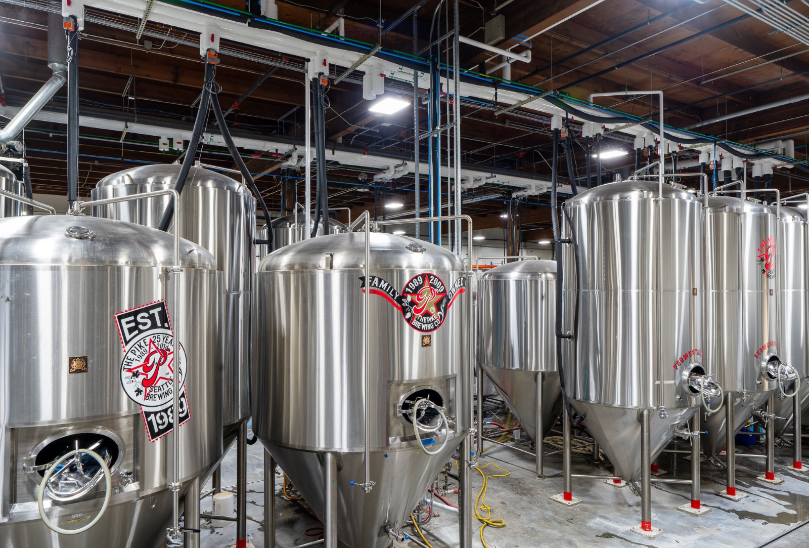 Large stainless steel brewing tanks with logos in an industrial facility with exposed ceiling pipes.