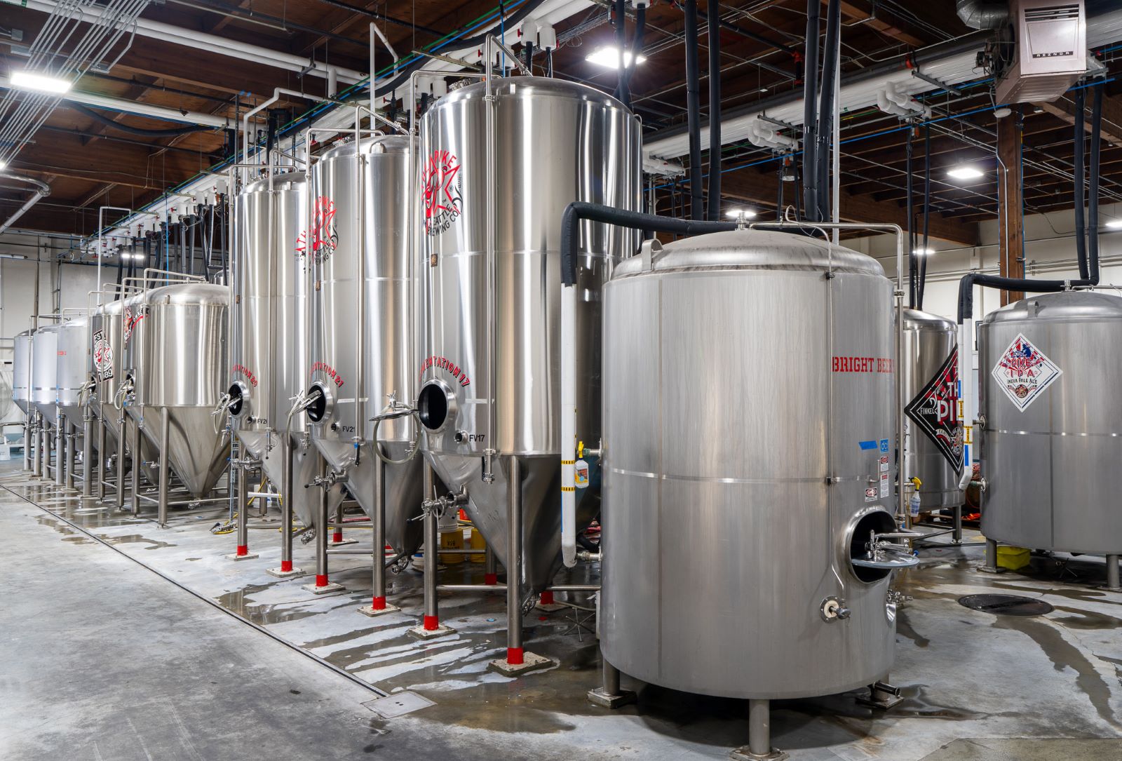 Row of stainless steel fermentation tanks in an industrial brewery with concrete floors and exposed ceilings.