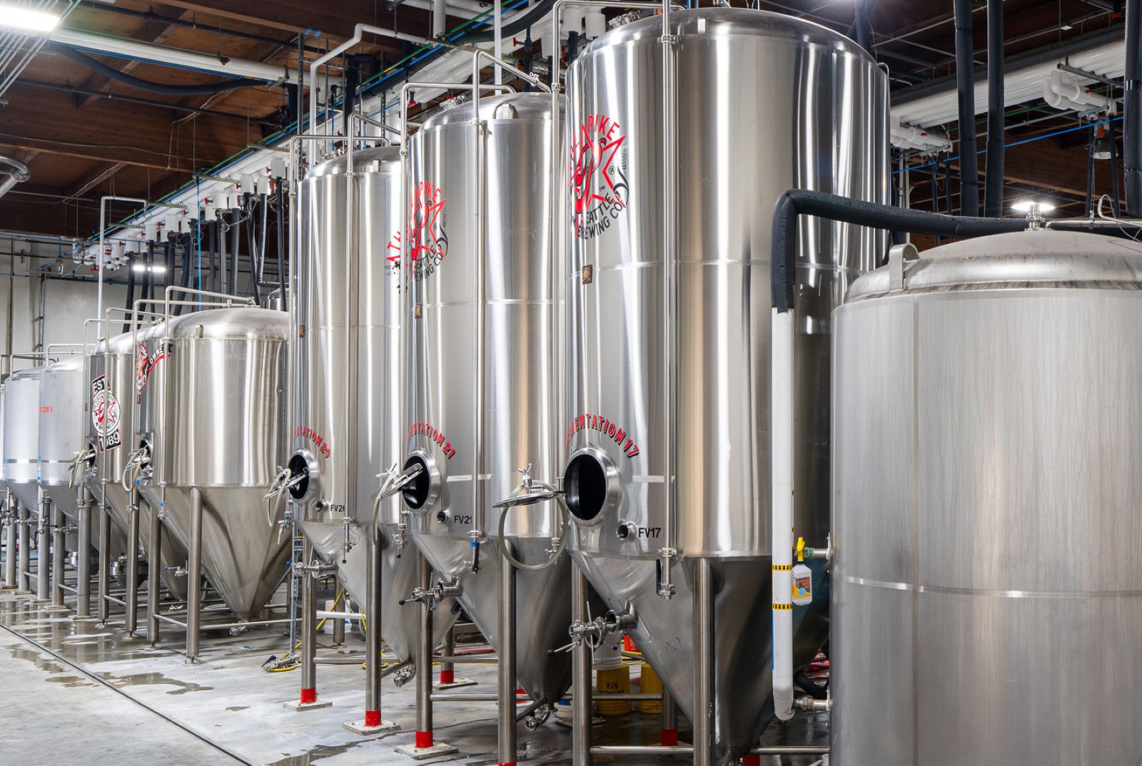 Row of stainless steel fermentation tanks in an industrial brewery with concrete floors and overhead pipes.