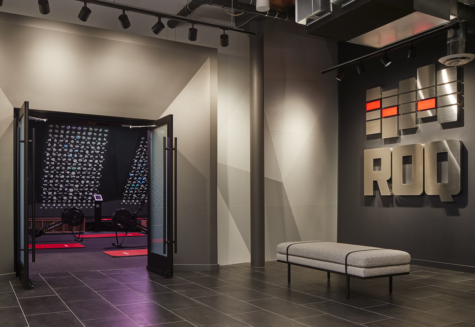 Modern gym lobby with 'ROQ' metal sign, grey bench, and open doors to a rowing and climbing workout room.