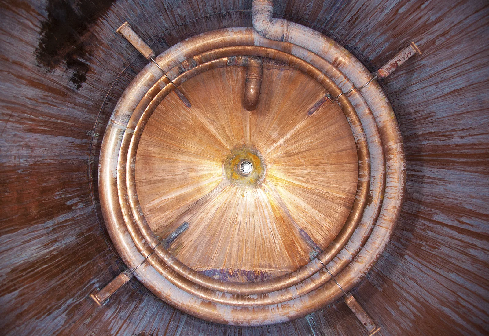 Interior view of a circular copper tank with concentric coils and a weathered, radial metallic texture.