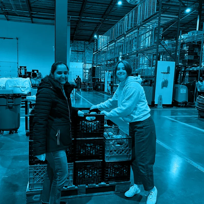 Two women smiling while sorting crates of produce in a large warehouse, with a blue color overlay.