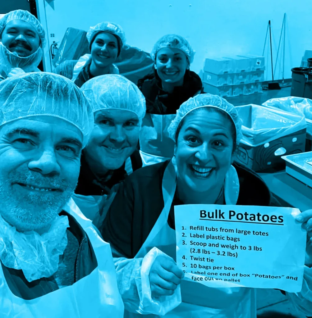 Smiling volunteers in hairnets and aprons hold a 'Bulk Potatoes' instruction sheet.