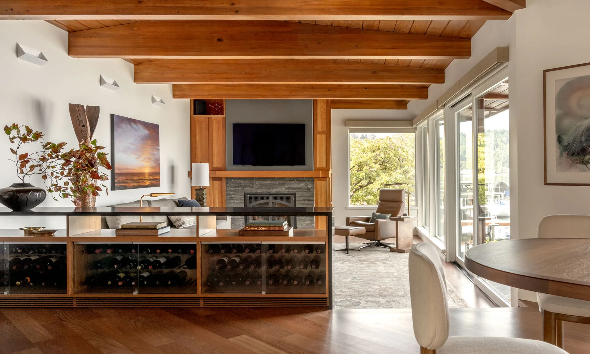 Warm living space featuring exposed wood ceiling beams, a built-in wine rack divider, and large windows.