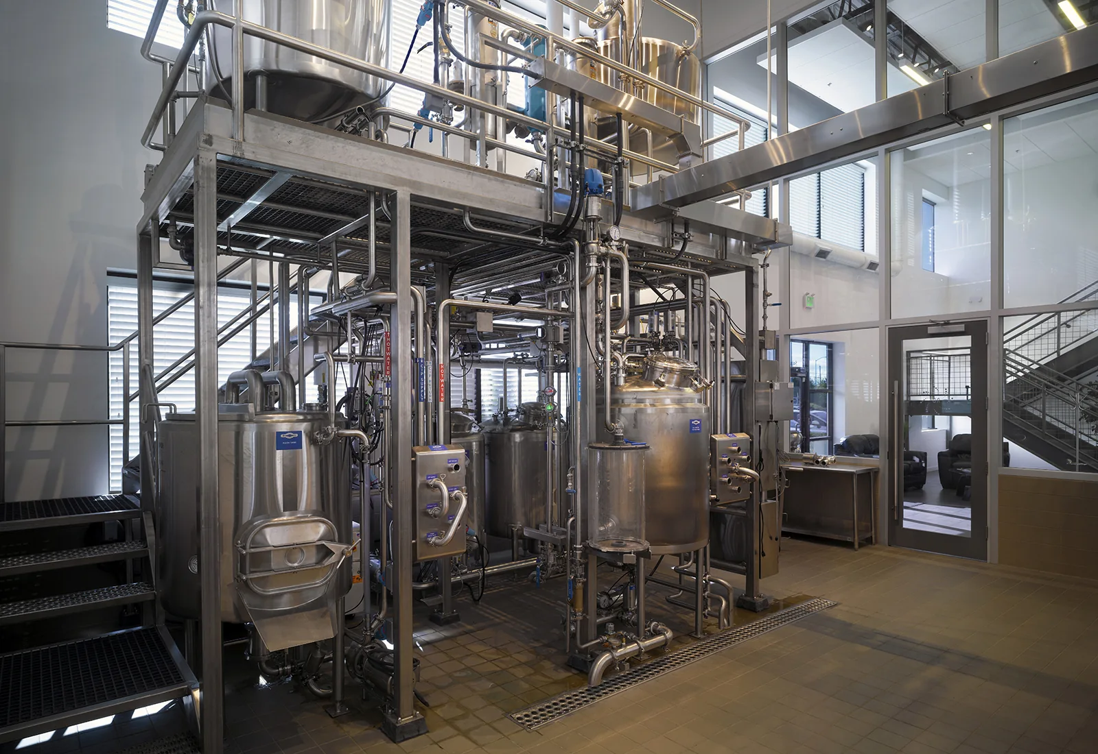 Industrial stainless steel brewing system in a modern facility with glass walls and tiled floors.
