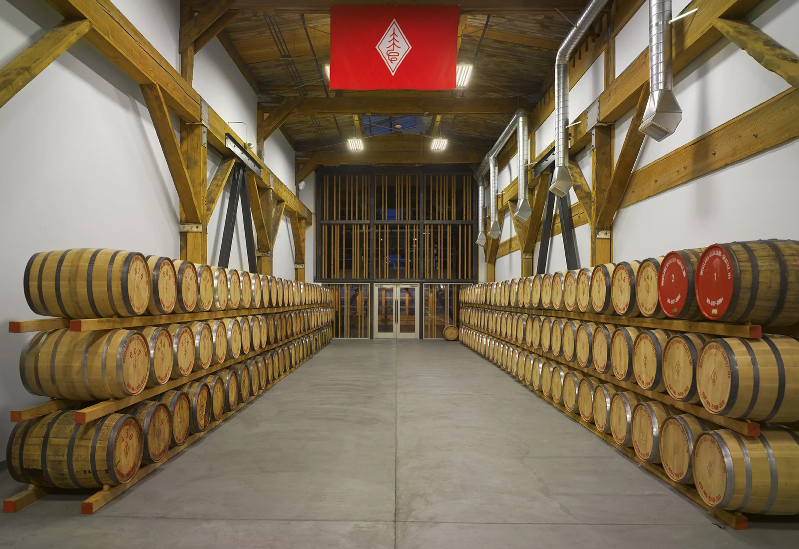 Wooden barrels stacked on racks in an industrial barrel room with exposed timber framing and a concrete floor.