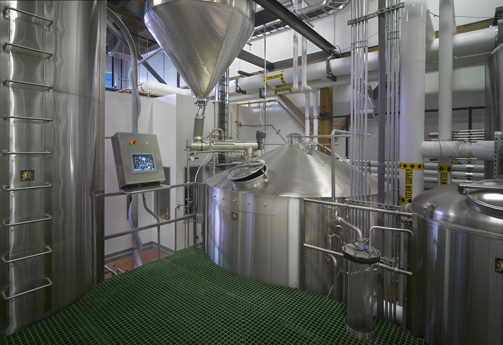 Industrial distillery interior featuring stainless steel mash tuns, overhead piping, and green grated flooring.
