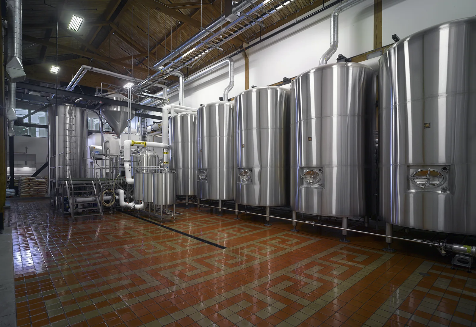 Stainless steel brewing tanks in an industrial mash room with patterned tile flooring and exposed wood ceiling.