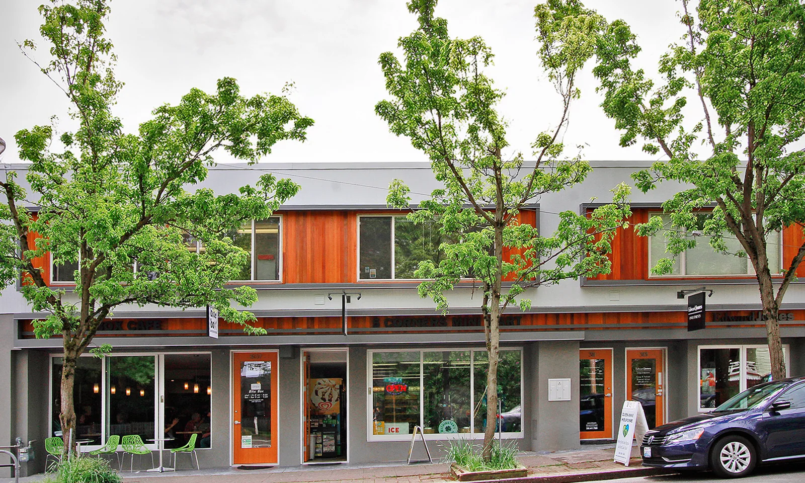 Modern mixed-use building with warm wood paneling, glass storefronts, and green street trees.