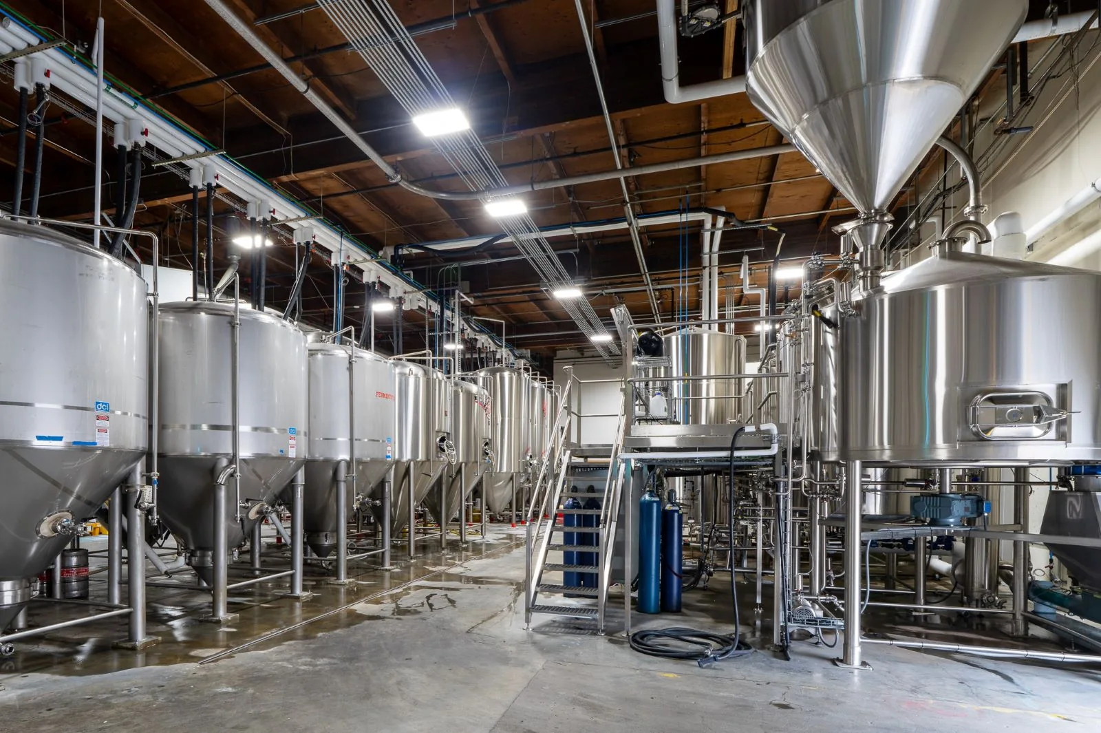 Industrial brewery interior featuring rows of stainless steel fermentation tanks, piping, and concrete floors.