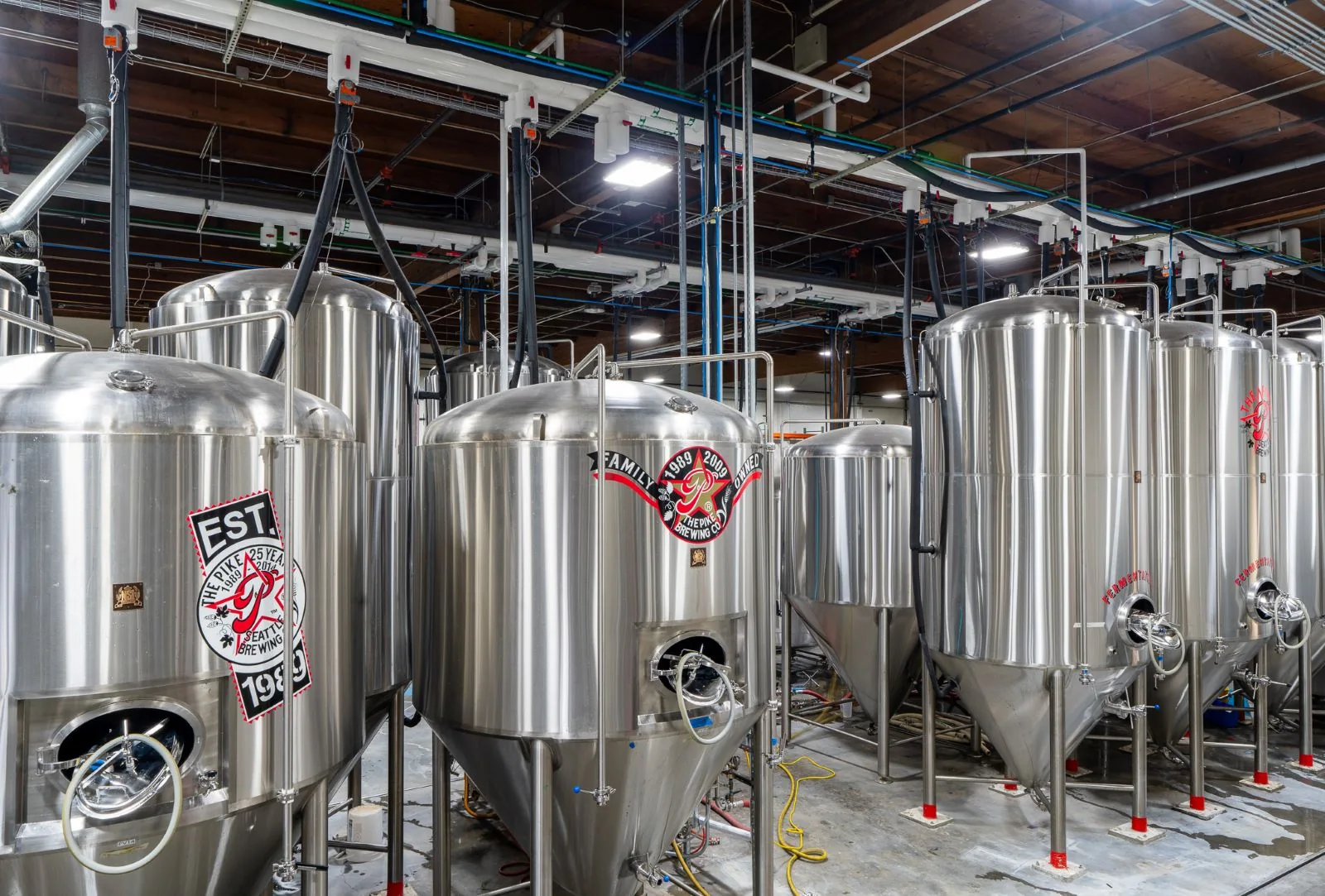Large stainless steel brewing tanks with logos in an industrial facility with exposed ceiling pipes.