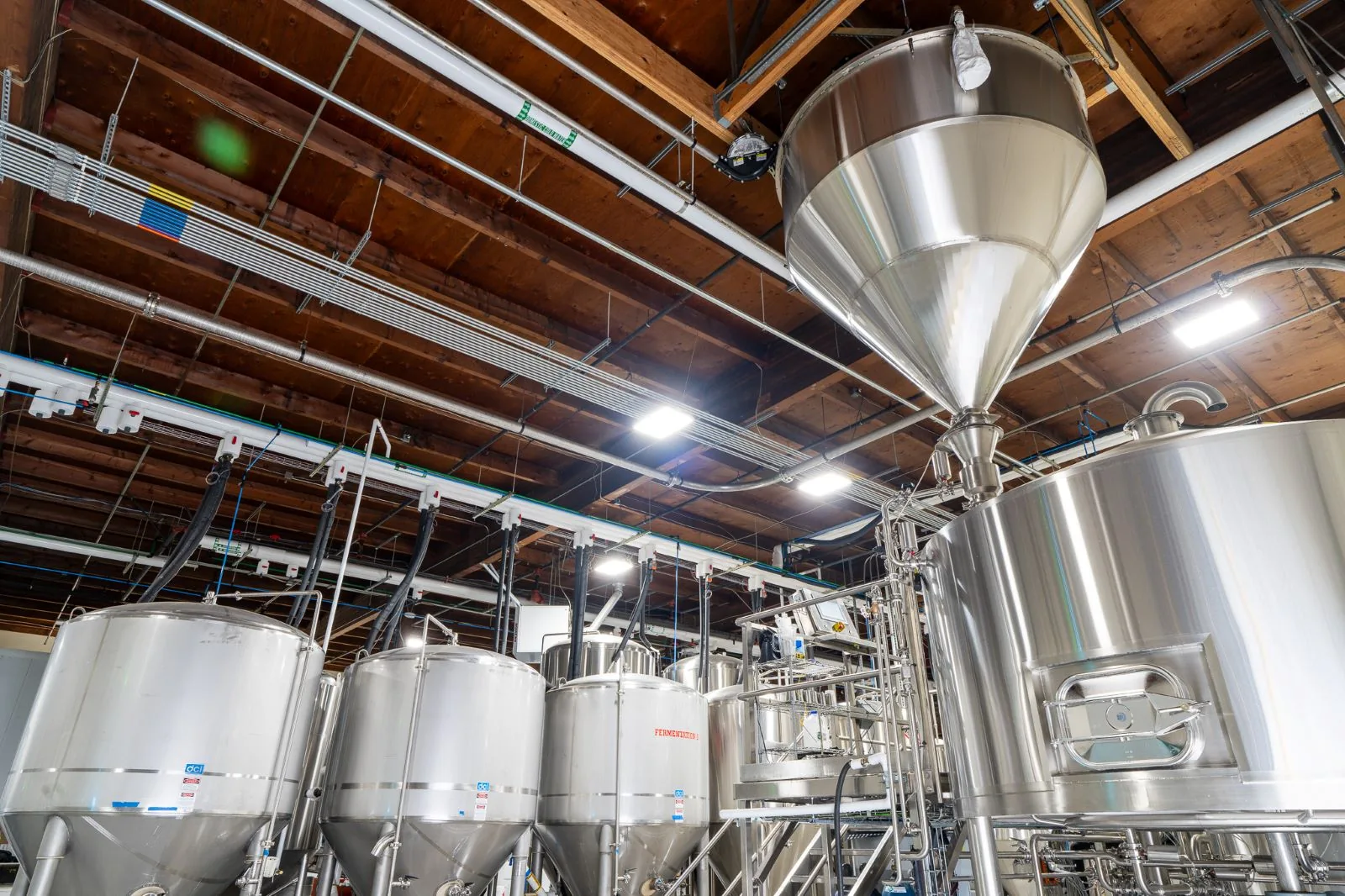 Stainless steel brewing tanks and equipment under an industrial exposed wood ceiling.