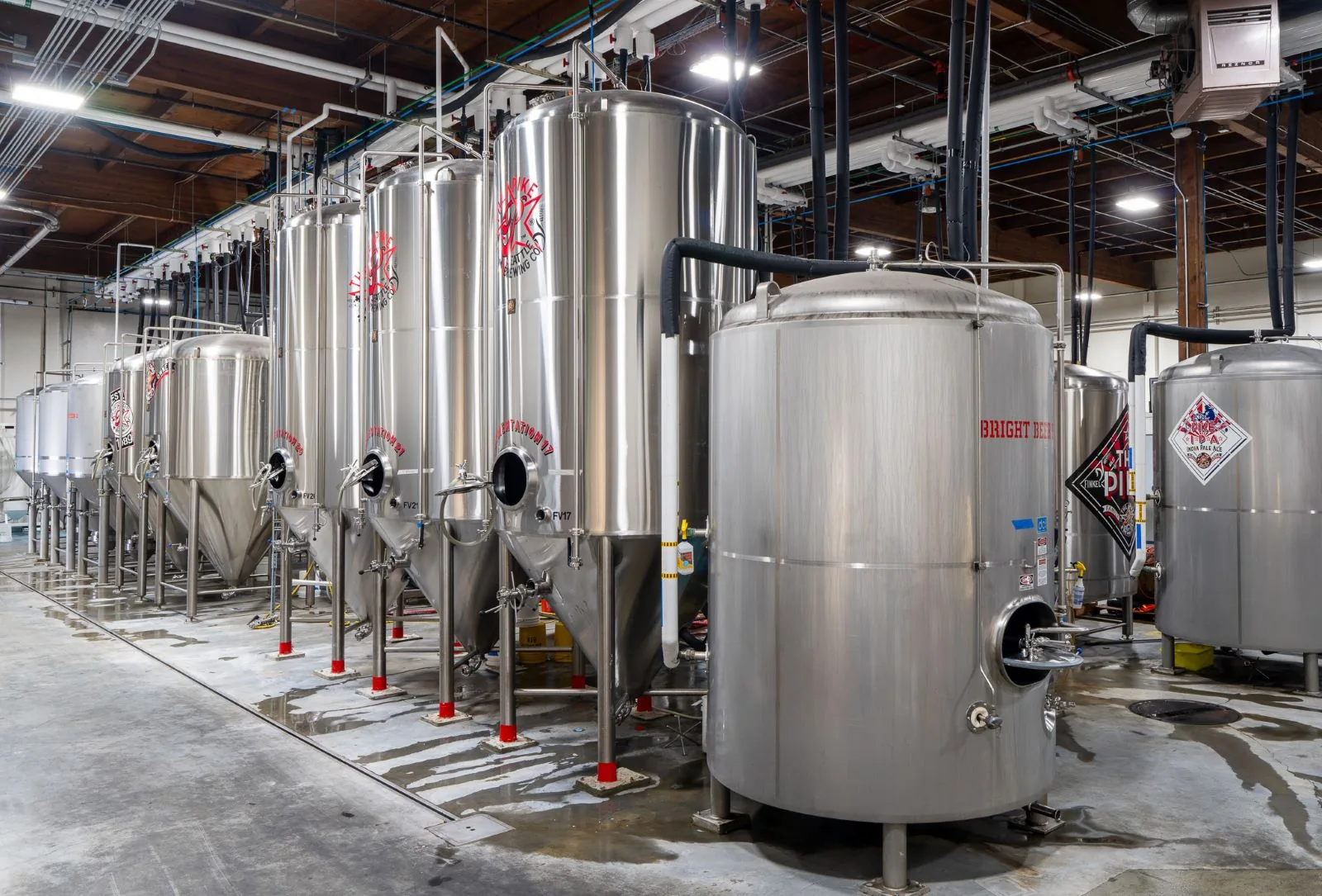 Row of stainless steel fermentation tanks in an industrial brewery with concrete floors and exposed ceilings.