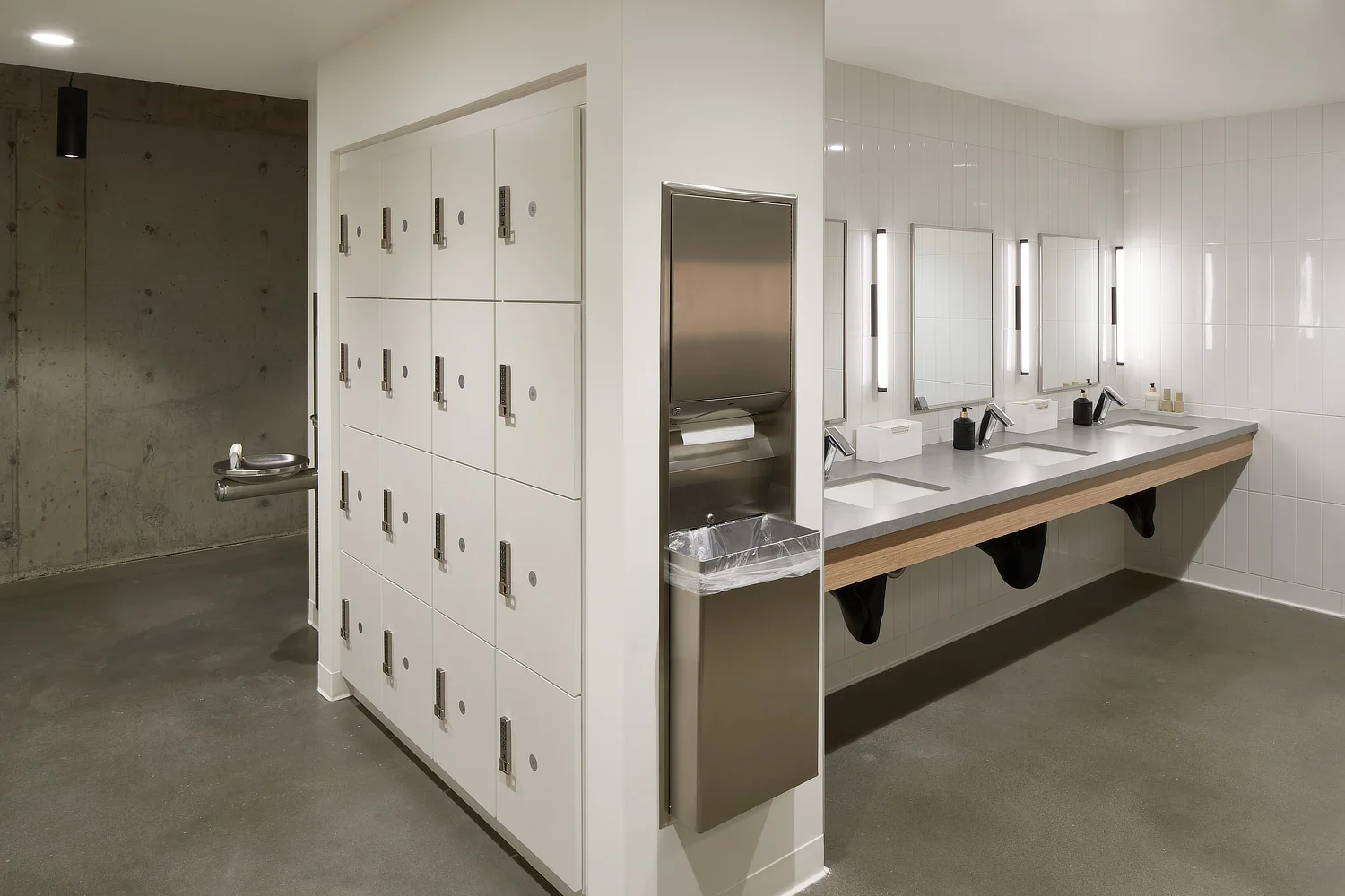 Modern restroom and locker area with white lockers, concrete floors, and a sleek vanity with vertical sconces.