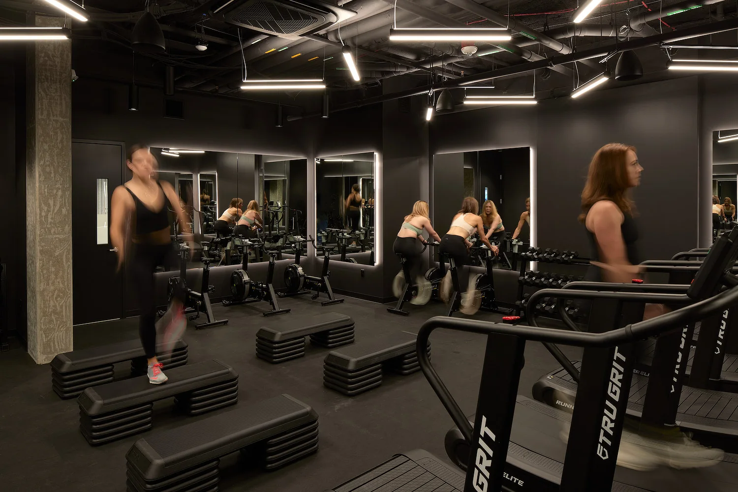 Sleek, dark-themed gym interior with linear LED lighting, mirrors, and people on treadmills and step platforms.
