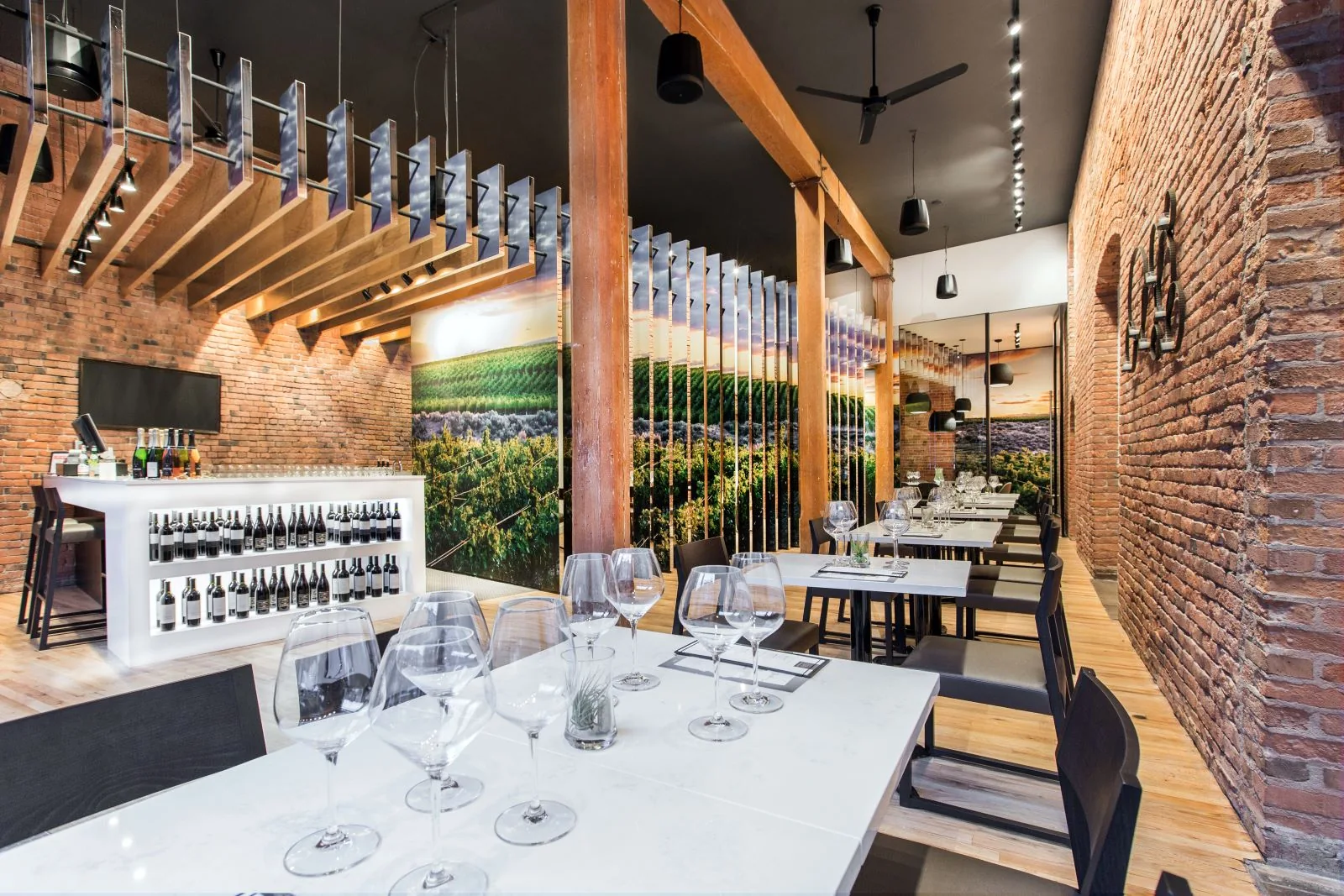 Modern tasting room with exposed brick walls, wooden columns, white tables with wine glasses, and a glowing bar.