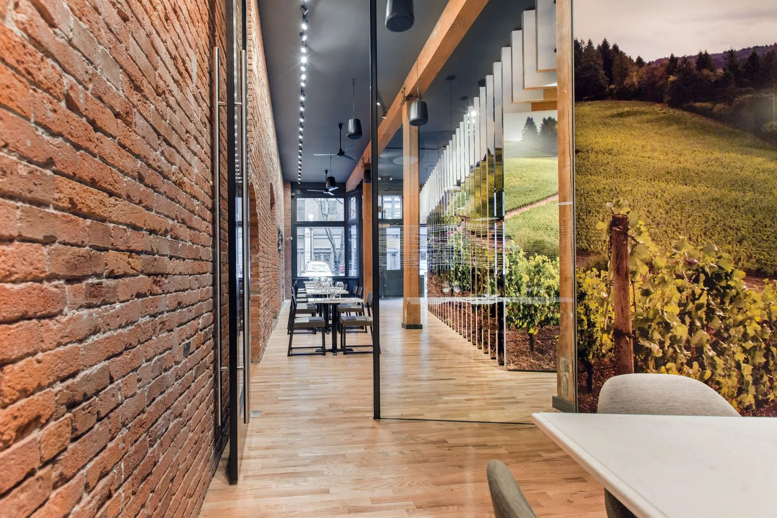 Modern tasting room interior with exposed brick, light wood floors, glass walls, and a vineyard mural.