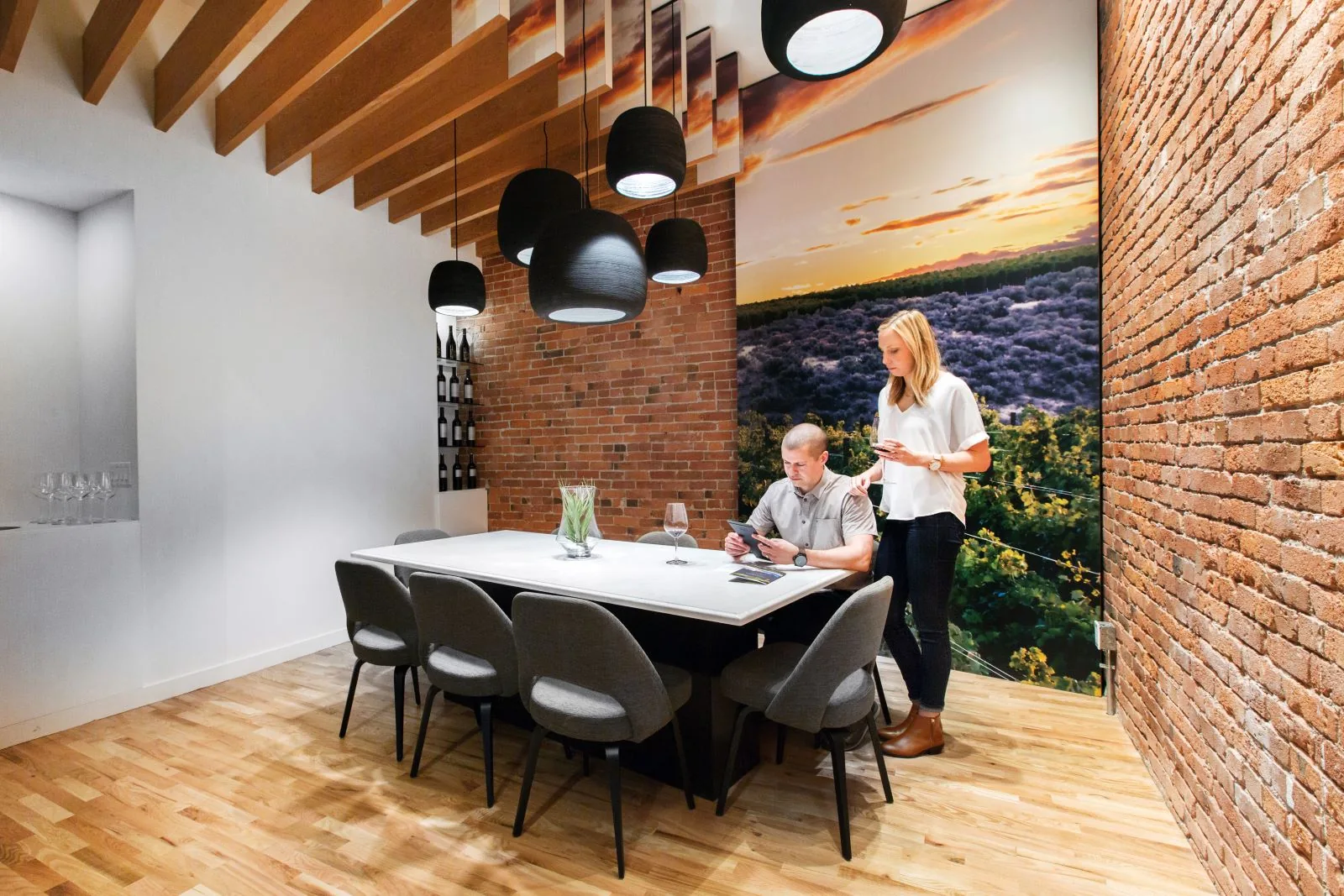 Two people in a modern tasting room with exposed brick walls, wood ceiling beams, and a large vineyard mural.
