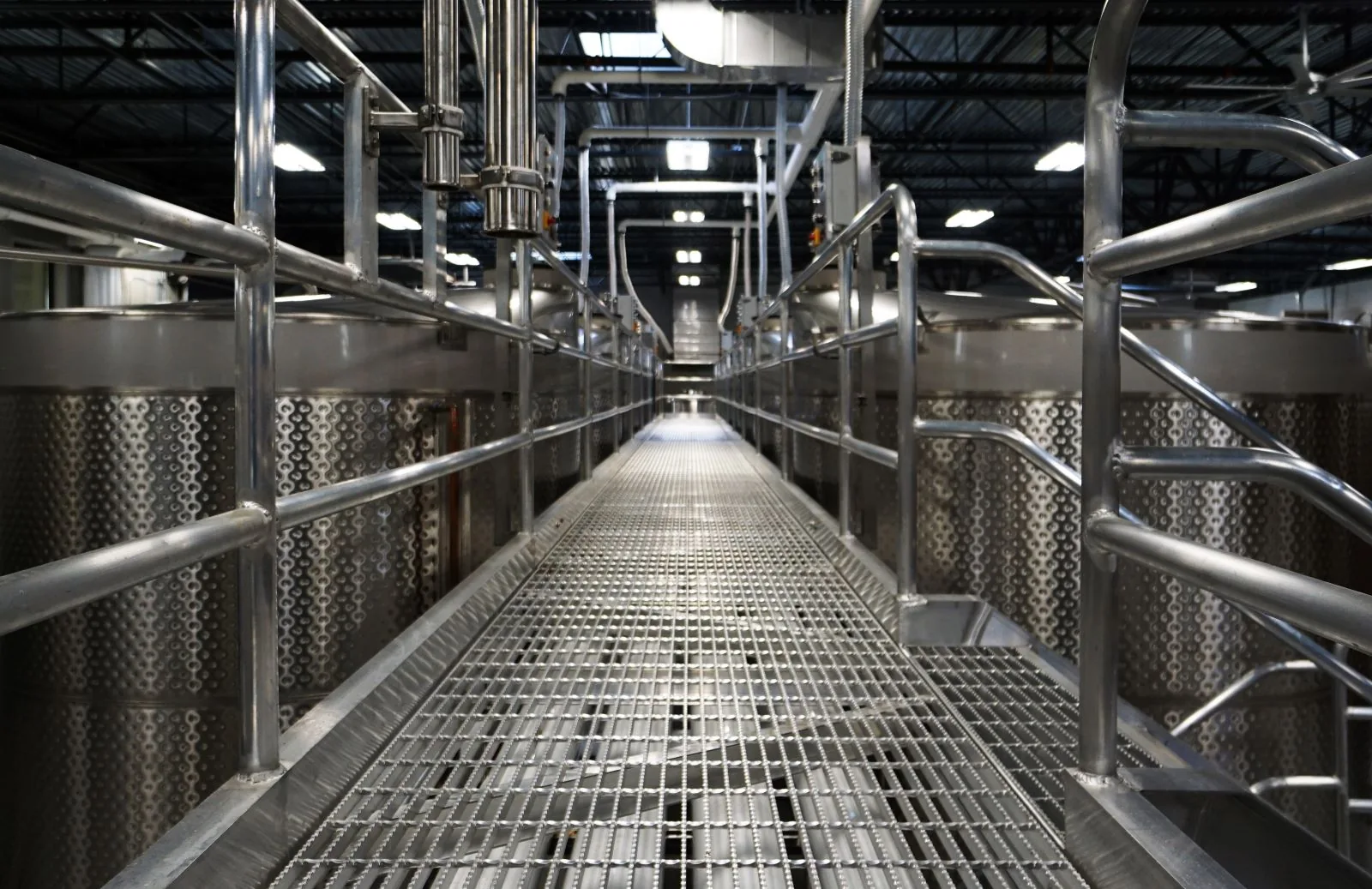 Industrial metal catwalk flanked by large stainless steel fermentation tanks in a modern brewery.