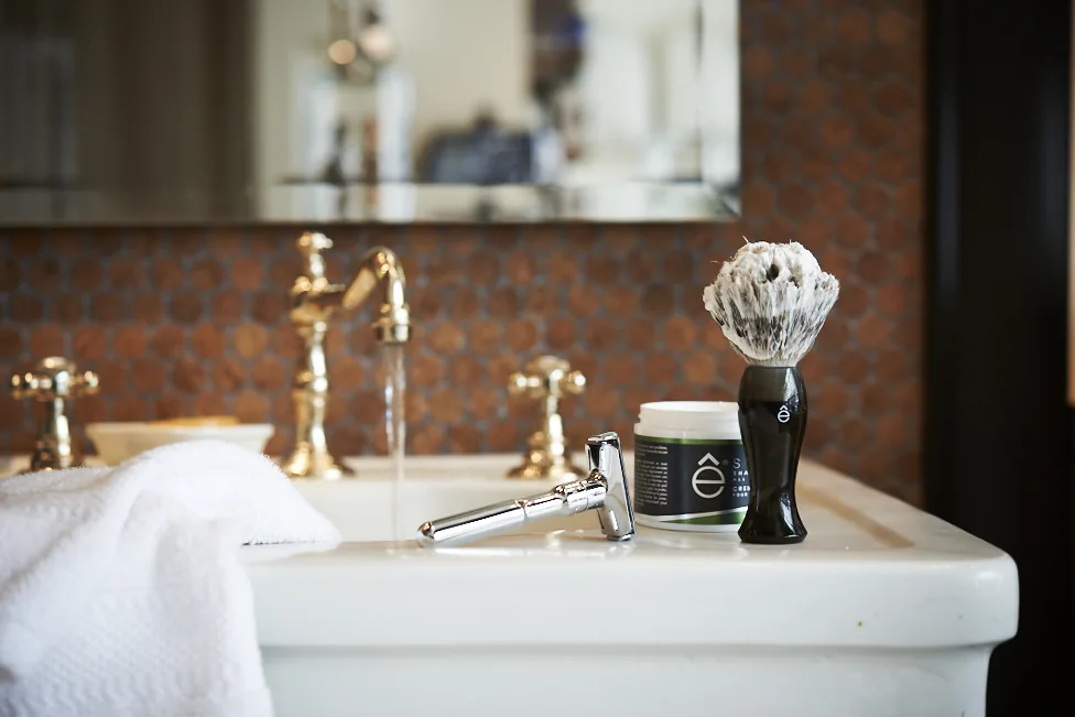 Classic shaving set with a razor and brush on a white sink with a running brass faucet and brown penny tile.