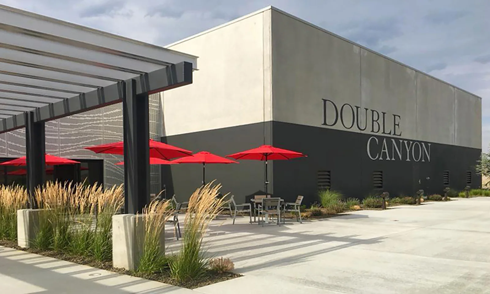 Modern concrete exterior of Double Canyon winery with red patio umbrellas, outdoor seating, and ornamental grasses.