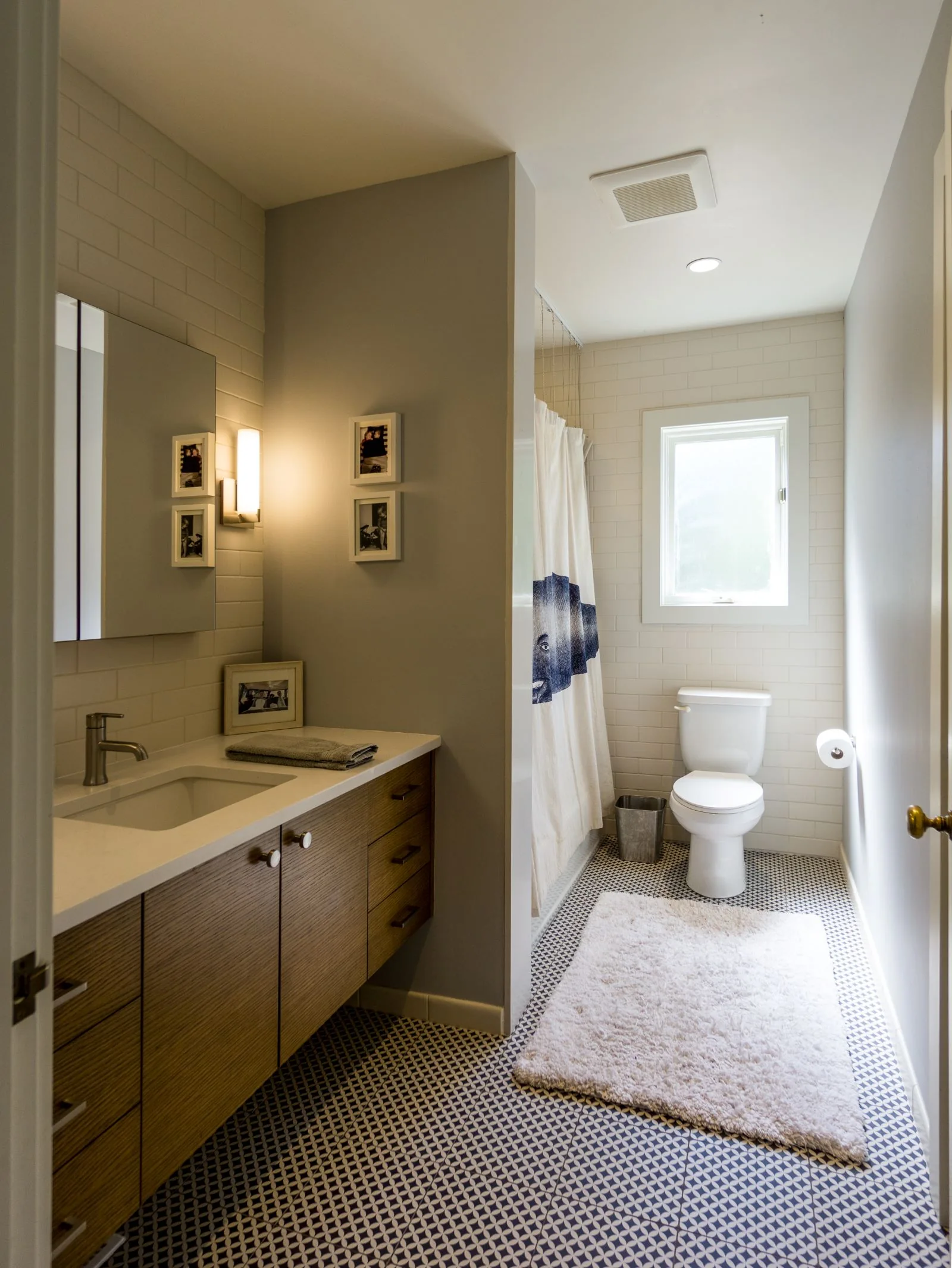 Modern bathroom with a warm wood vanity, patterned tile floor, and a toilet next to a small window.