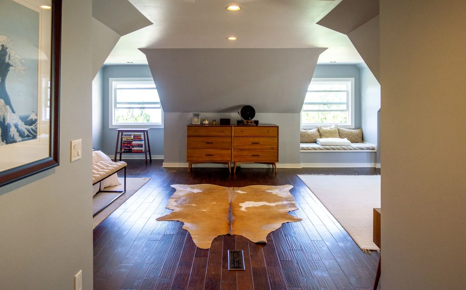 Bright attic bedroom featuring dark wood floors, a cowhide rug, mid-century dresser, and cozy window seat.