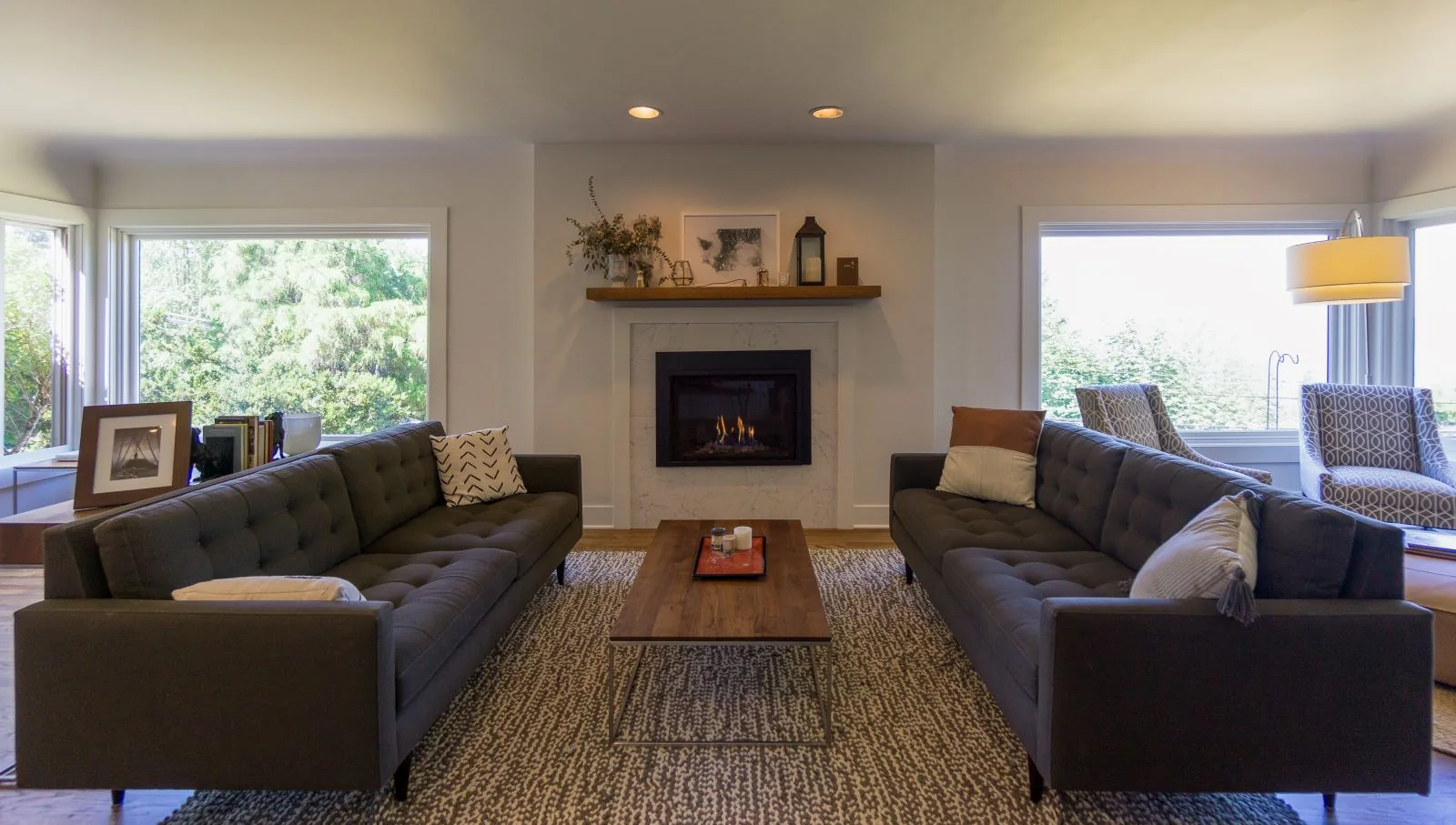 Modern living room with two grey tufted sofas facing a fireplace, a wooden coffee table, and large windows.