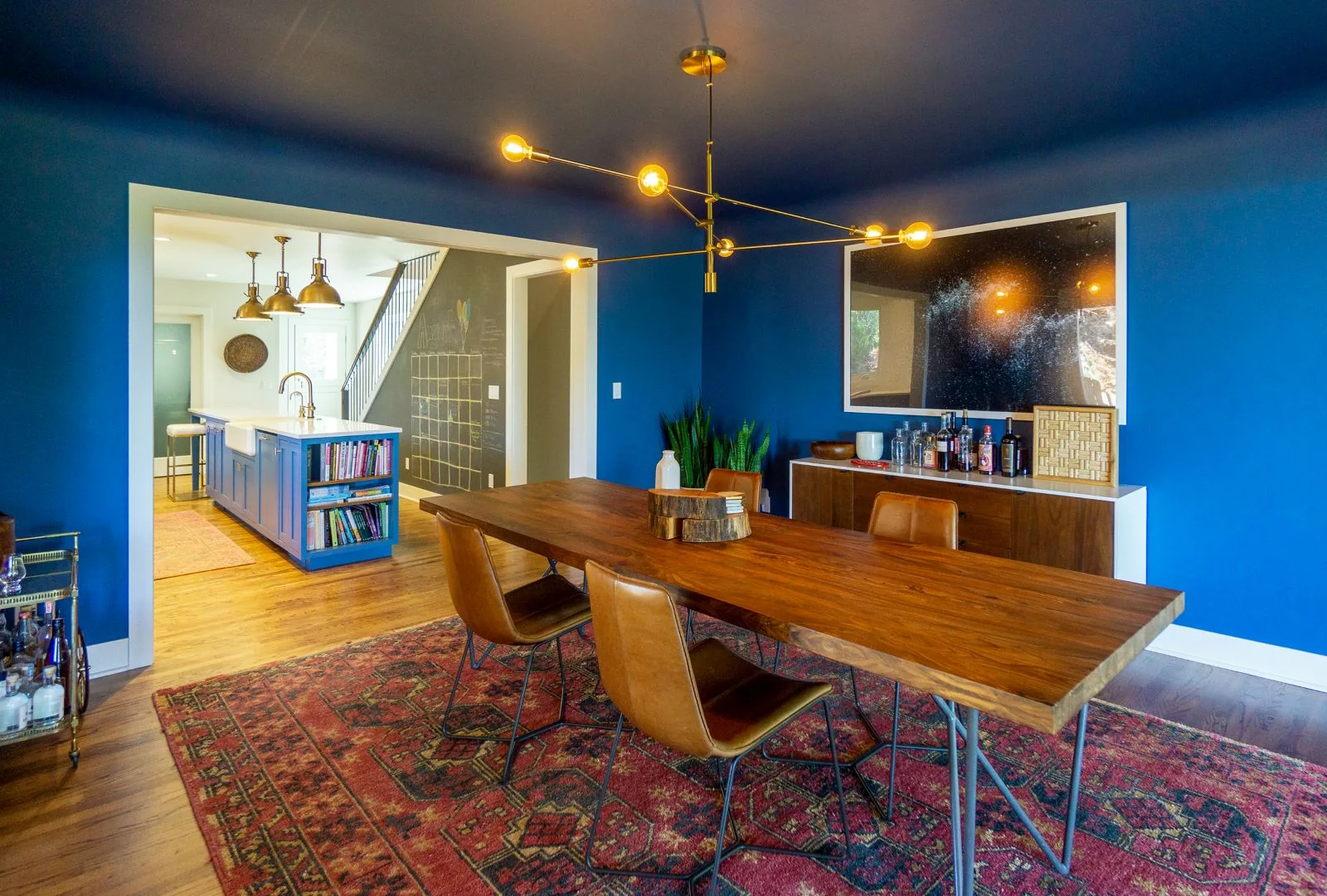 Bold blue dining room with a wooden table, leather chairs, a red patterned rug, and a modern brass chandelier.