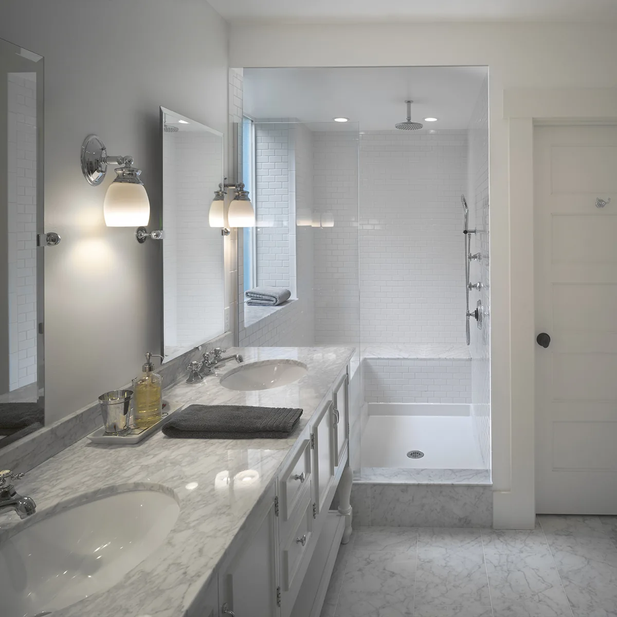 Classic white bathroom with a marble double vanity, sconce lighting, and a glass-enclosed subway tile shower.