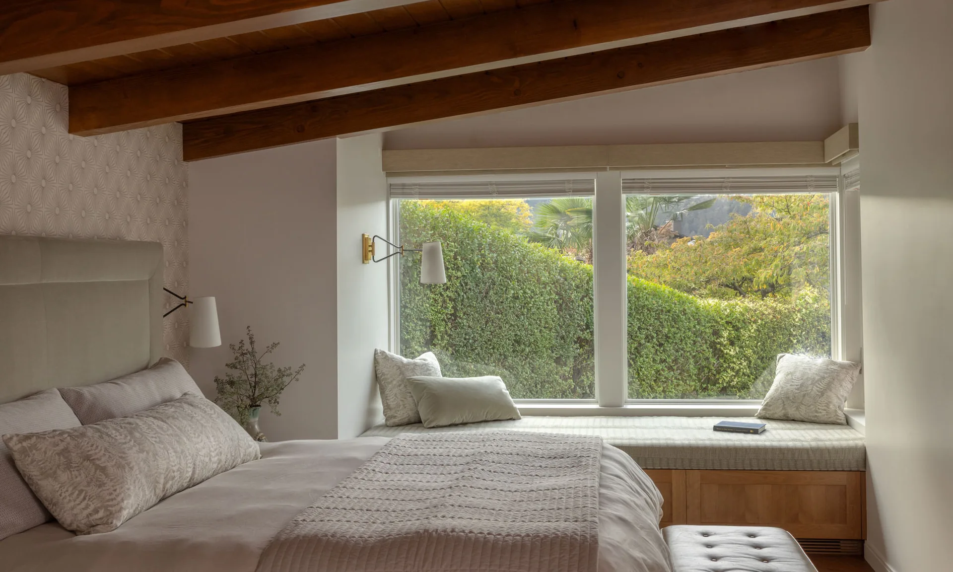 Bedroom with exposed wood beams, neutral bedding, and a cozy window seat looking out at lush green hedges.