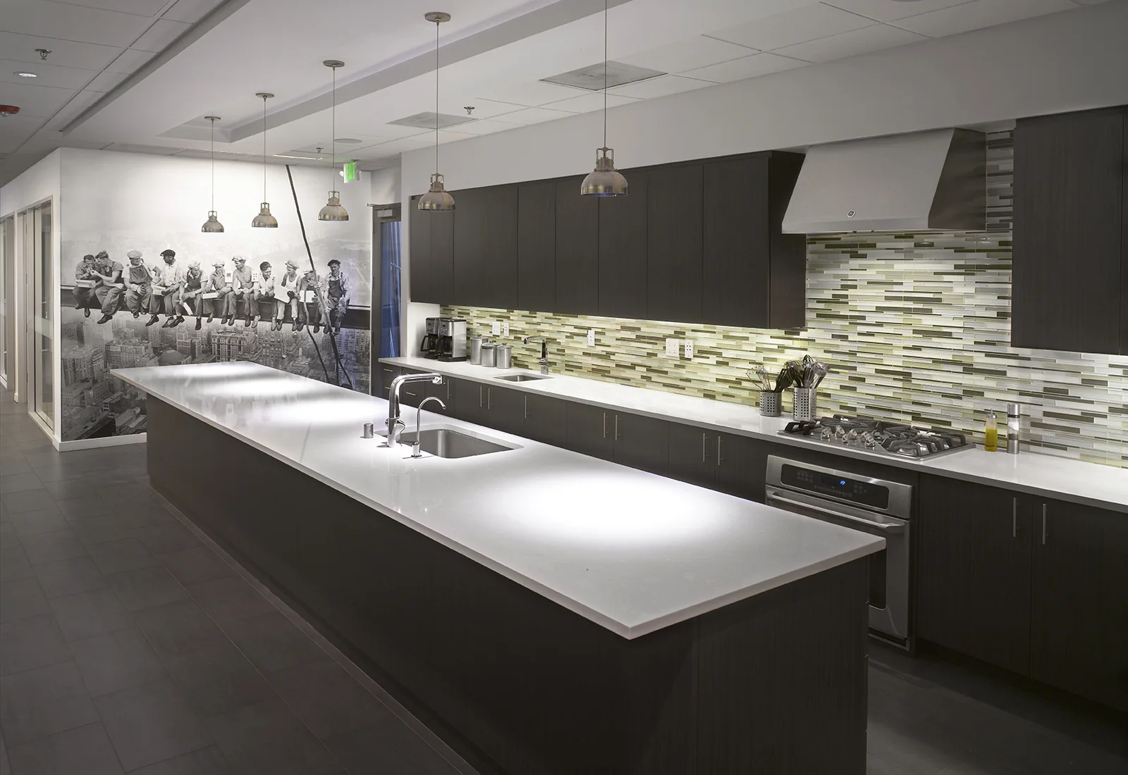 Modern office kitchen with dark wood cabinets, a long white island, and a historic construction worker wall mural.
