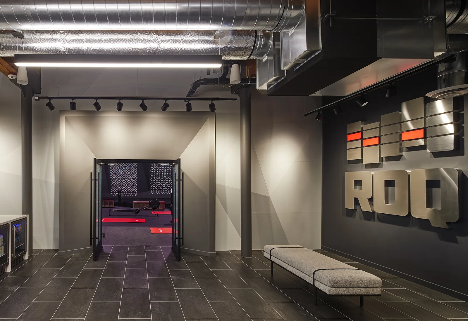 Modern industrial gym lobby with a grey bench, dark tiled floors, and a large metal ROQ sign on a black wall.
