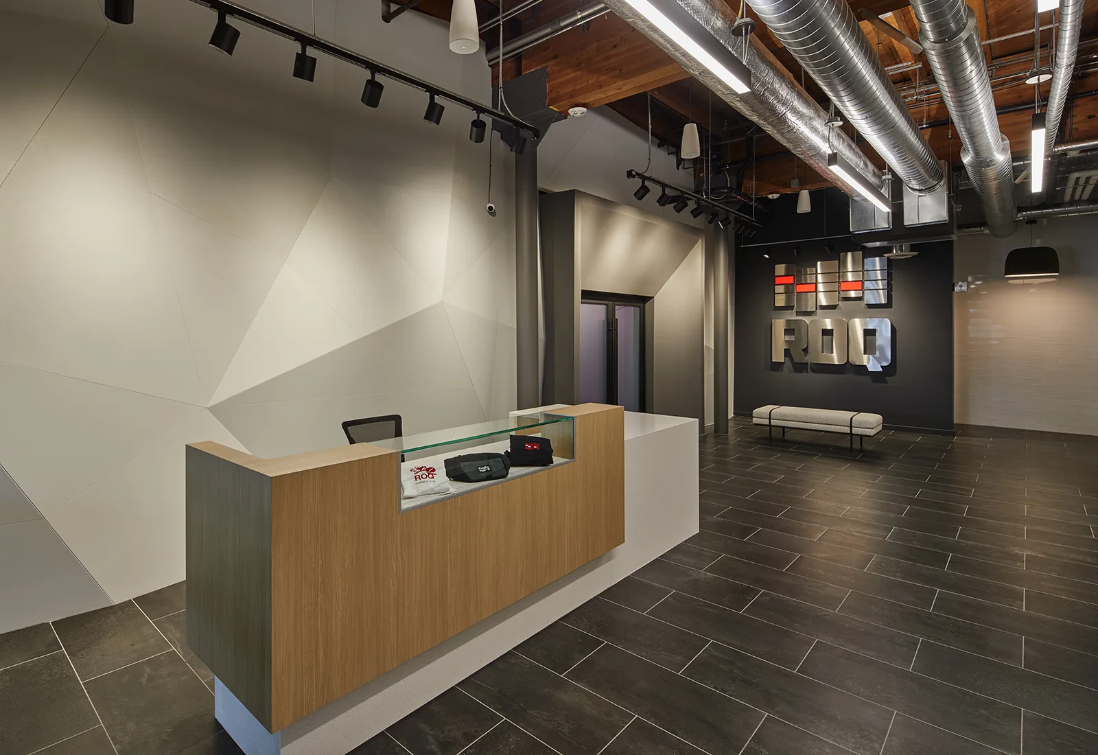 Modern lobby with a wood reception desk, geometric accent wall, dark tile floors, and industrial exposed ceiling.