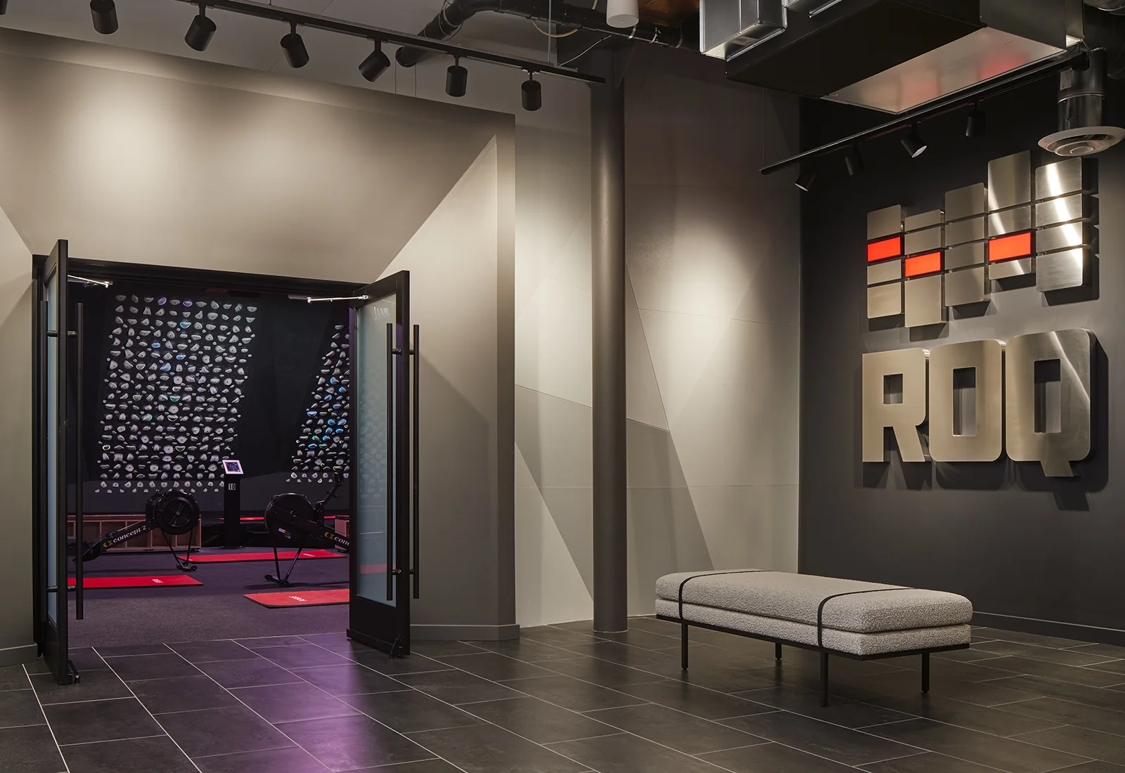Modern gym lobby with 'ROQ' metal sign, grey bench, and open doors to a rowing and climbing workout room.