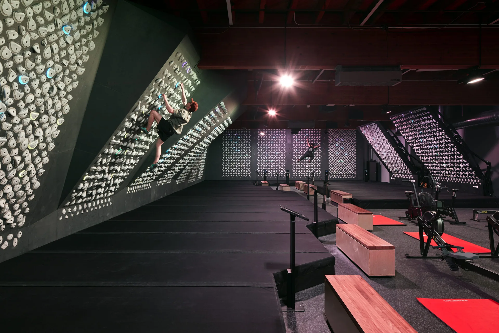 Climber on an overhanging wall inside a modern, industrial climbing gym with fitness equipment and dark mats.