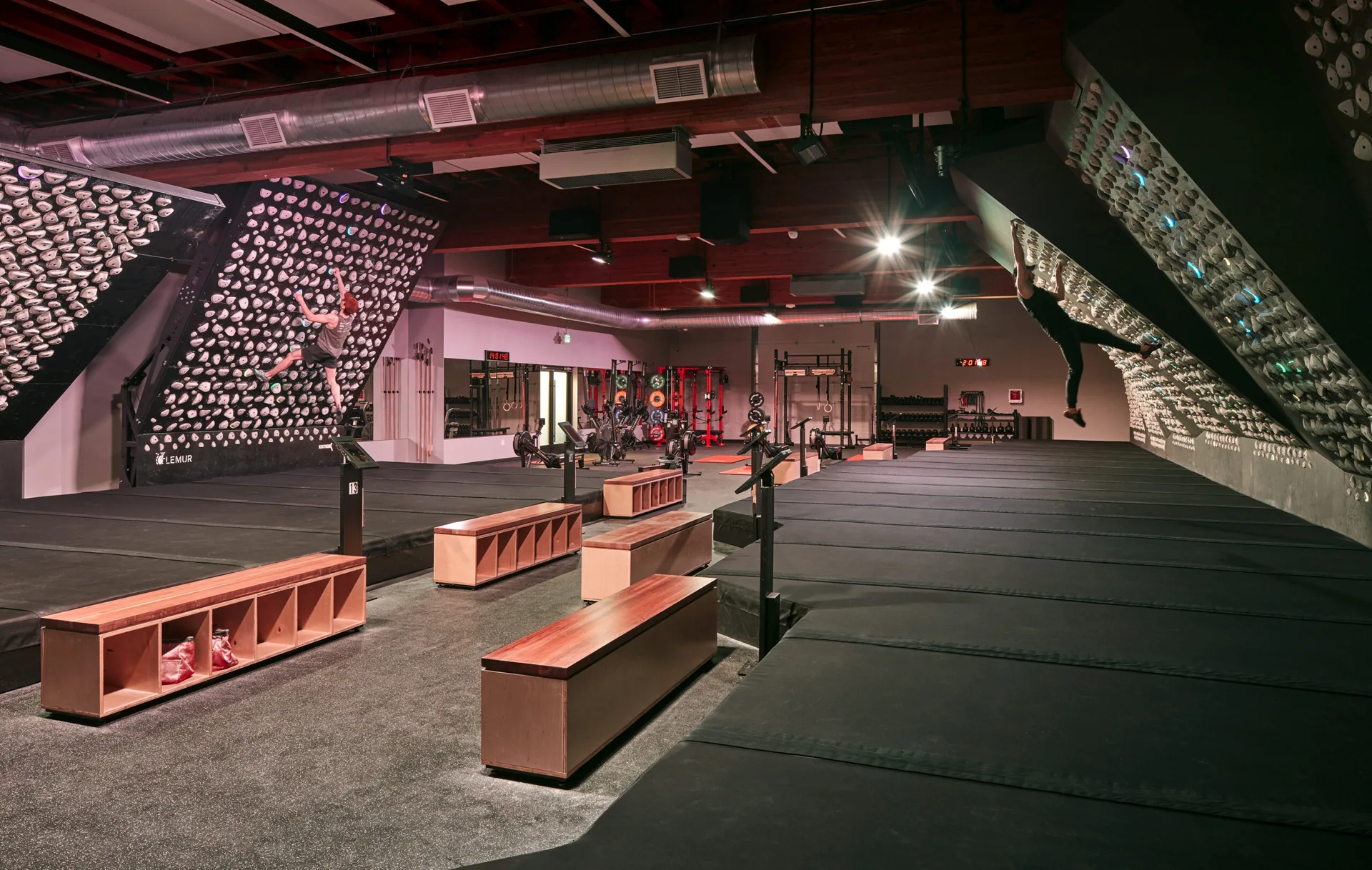 Climbers on angled bouldering walls in a modern training gym with wooden benches and black padded flooring.